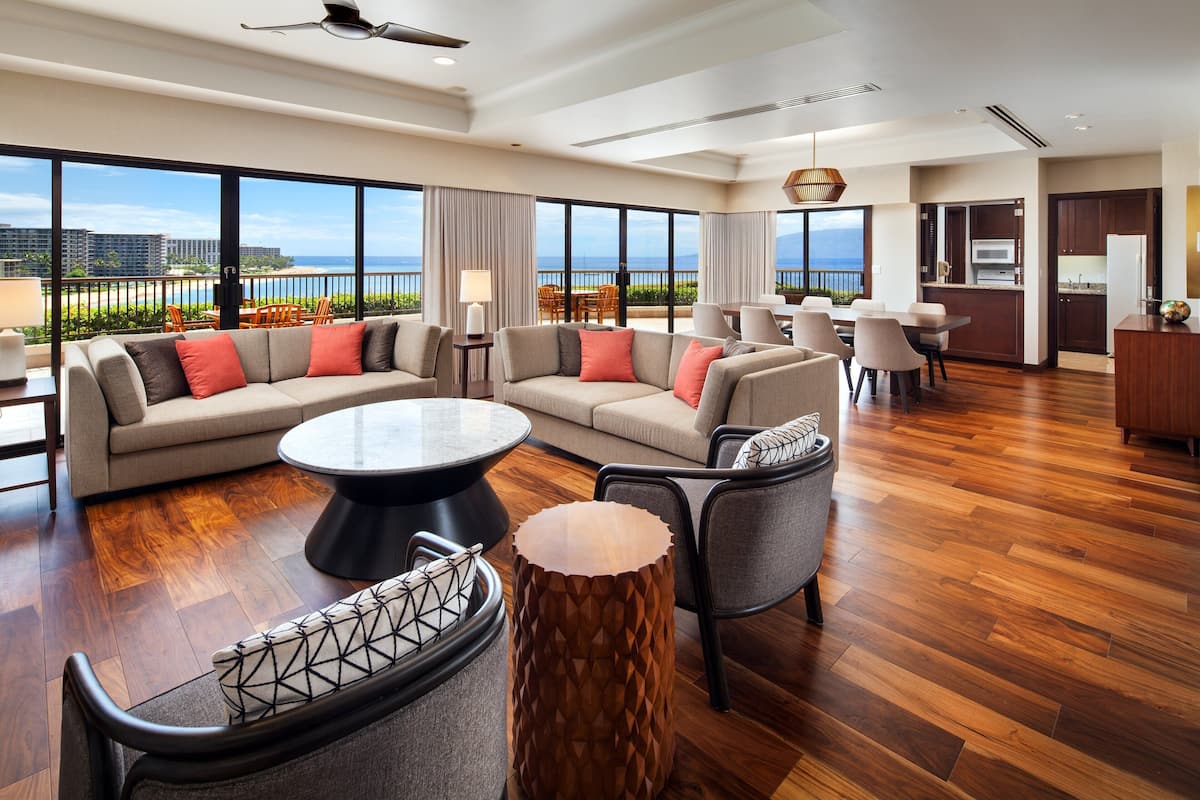 A spacious living area with a large sofa, a coffee table, and a dining table. There are multiple chairs and a kitchen area visible in the background.