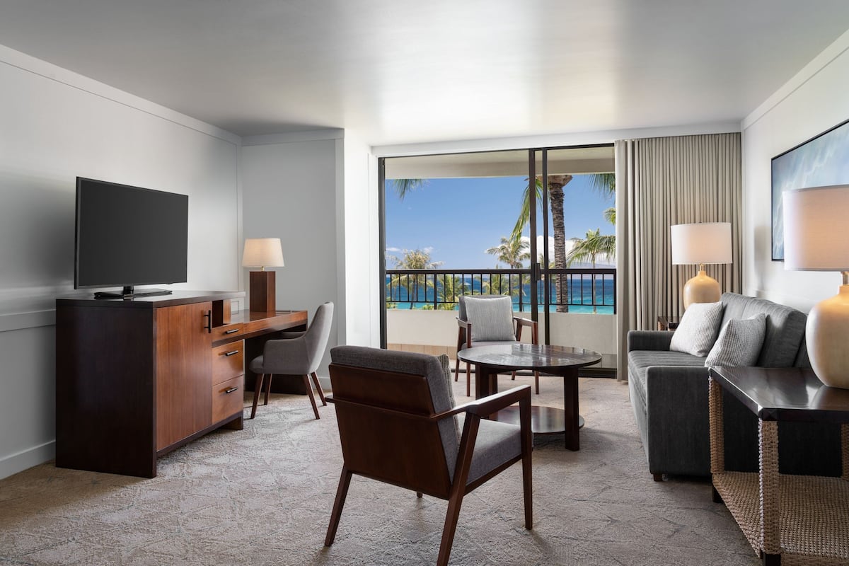A modern hotel room with a flat-screen TV, a wooden desk, a sofa, a dining table, and a balcony with a view of the ocean and palm trees.