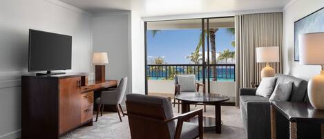 A modern hotel room with a flat-screen TV, a wooden desk, a sofa, a dining table, and a balcony with a view of the ocean and palm trees.