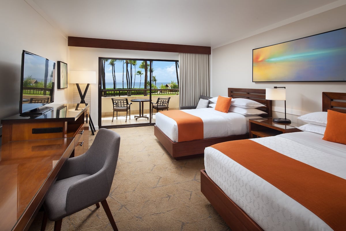 A hotel room with two beds, a desk, a chair, a TV, and a balcony with a view of palm trees and the ocean.