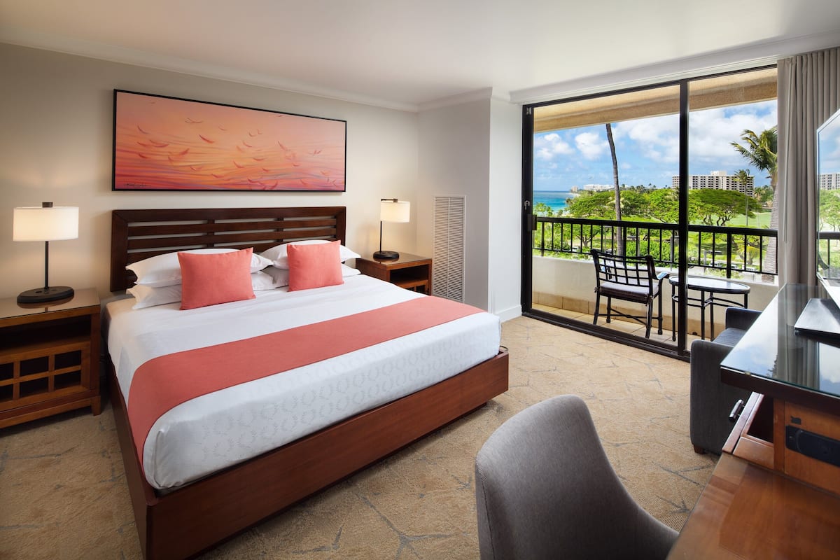 A hotel room with a large bed, two bedside tables, a desk, and a balcony with a view of the ocean and palm trees.