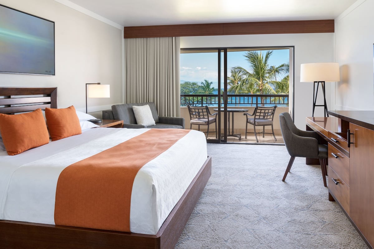 A hotel room with a large bed, a desk, a chair, a balcony with a view of palm trees and the ocean.