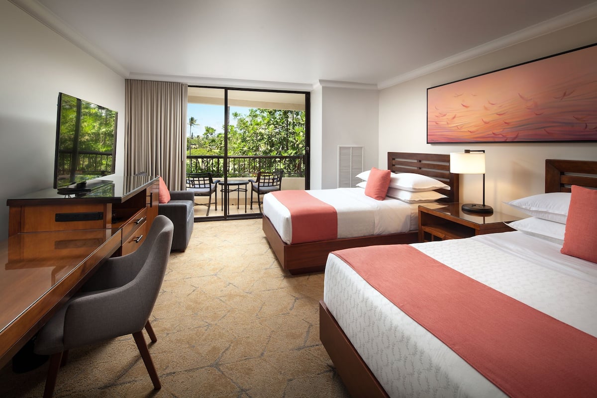 A hotel room with two beds, a desk, a chair, a TV, and a balcony with a view of trees.