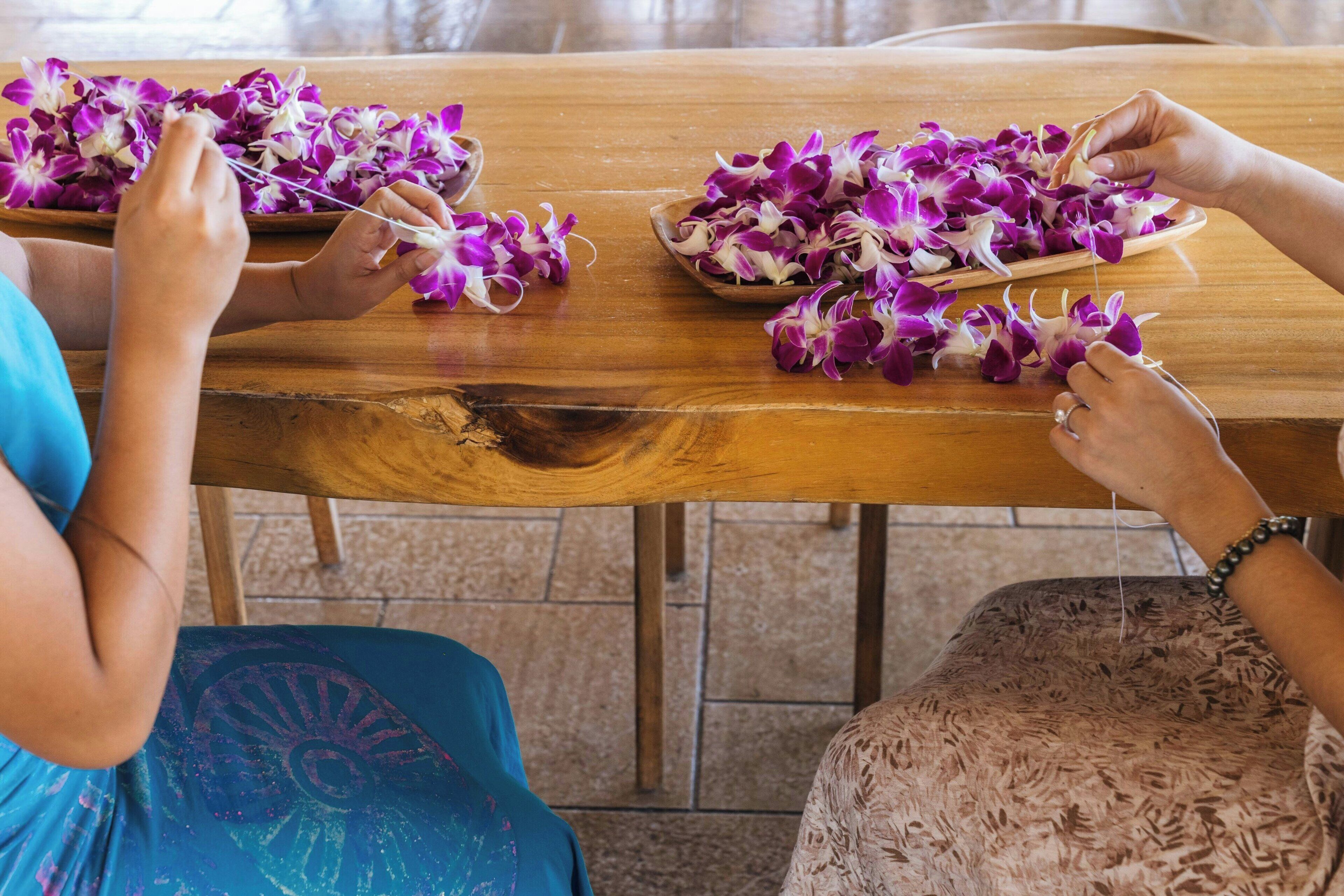 Una mesa de madera con flores moradas y blancas, y una persona ensartándolas con un hilo.
