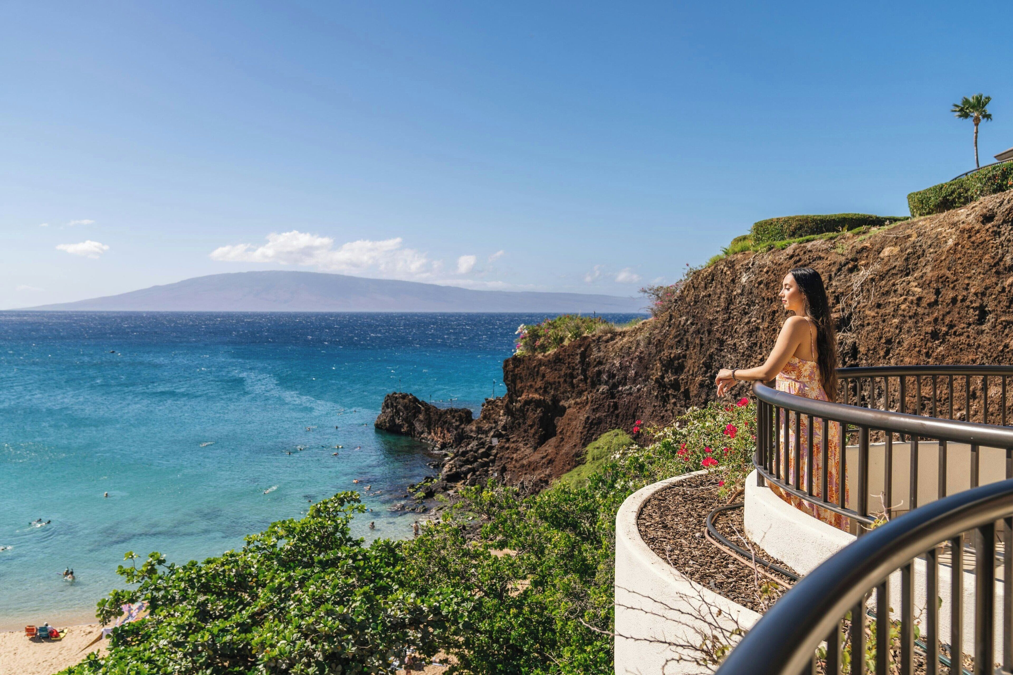 Une vue sur la côte, avec une personne sur un balcon donnant sur la mer et une montagne à l’horizon.