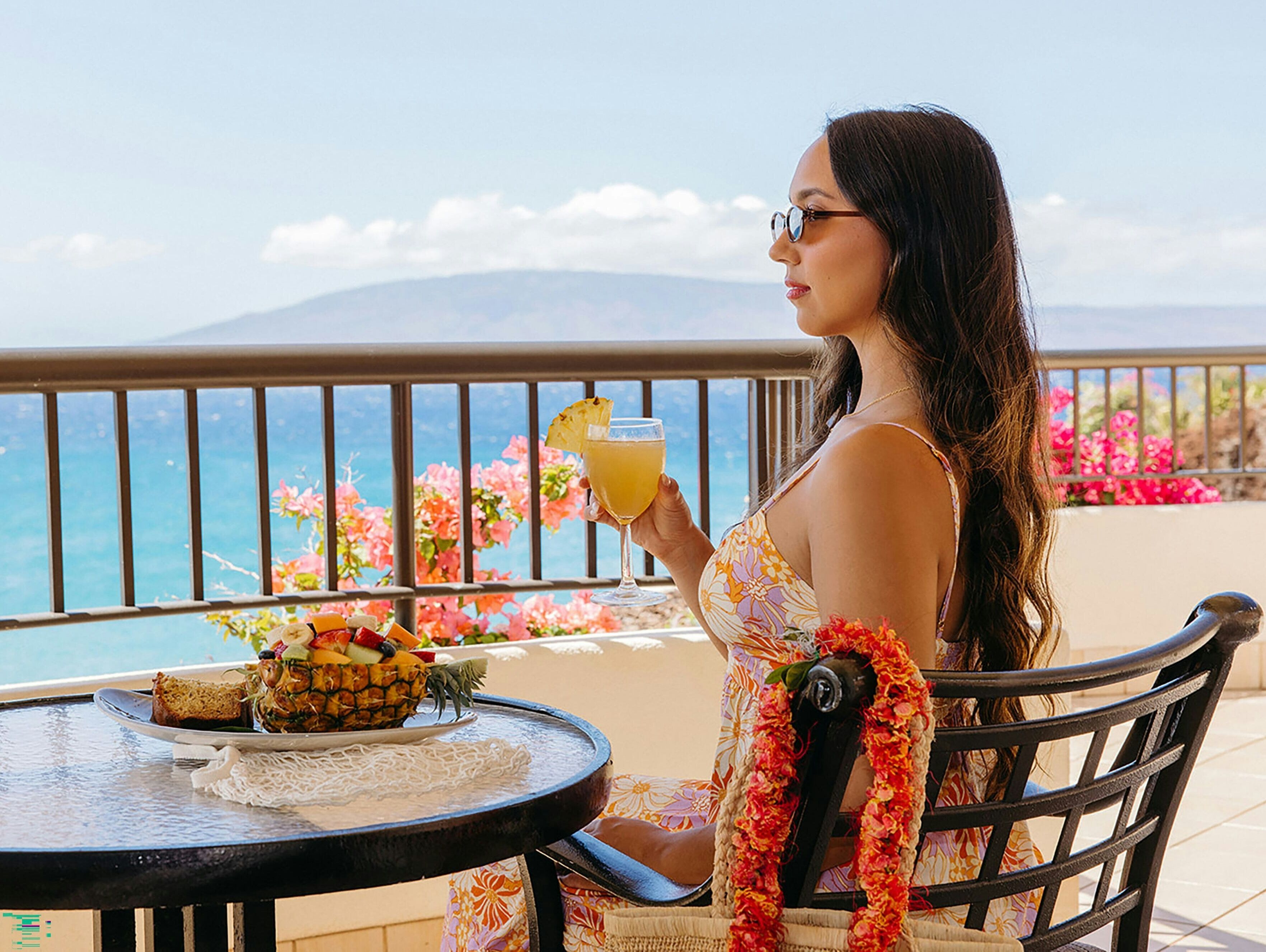Una mujer sentada a una mesa con una bebida y un frutero, con vistas a un paisaje con flores y una barandilla.