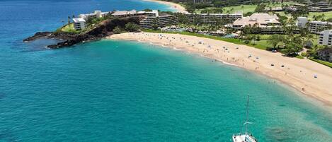An aerial view of a coastal resort with a beach, clear turquoise waters, and a yacht anchored near the shore.