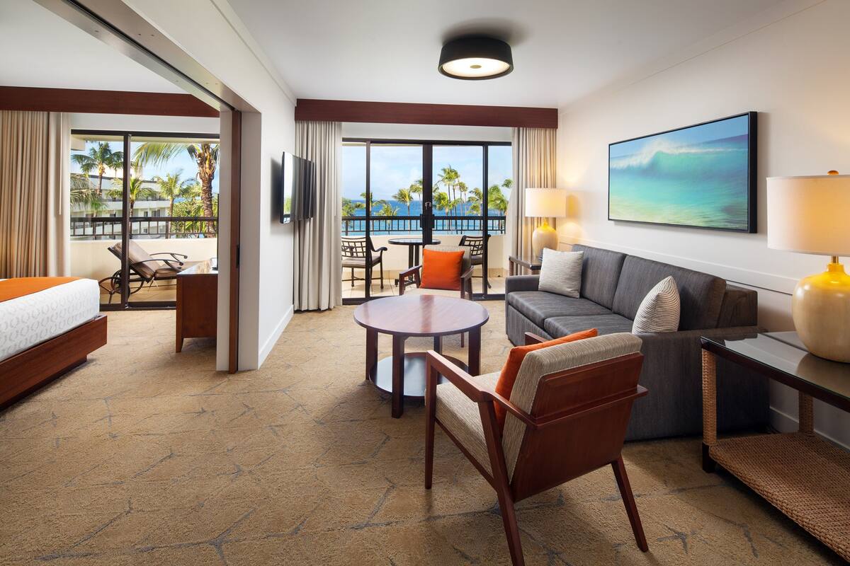 A hotel room with a bed, a sofa, a dining table, and a balcony with a view of palm trees and the ocean.