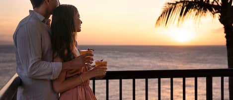 Una pareja está de pie en un balcón con vista a un atardecer y el océano al fondo.