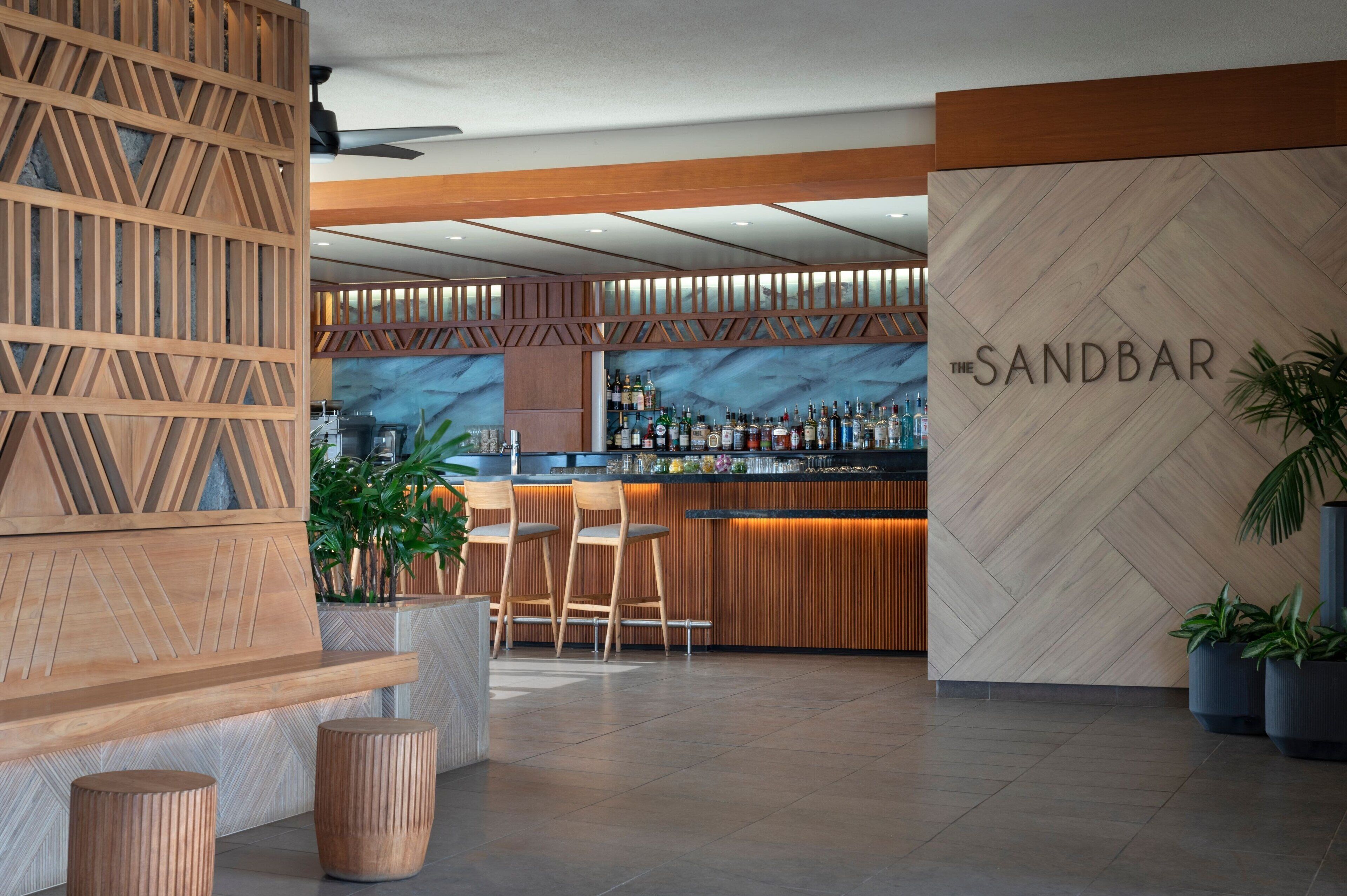 A modern bar area with a wooden partition, bar stools, and a wall with the name 'The Sandbar'.