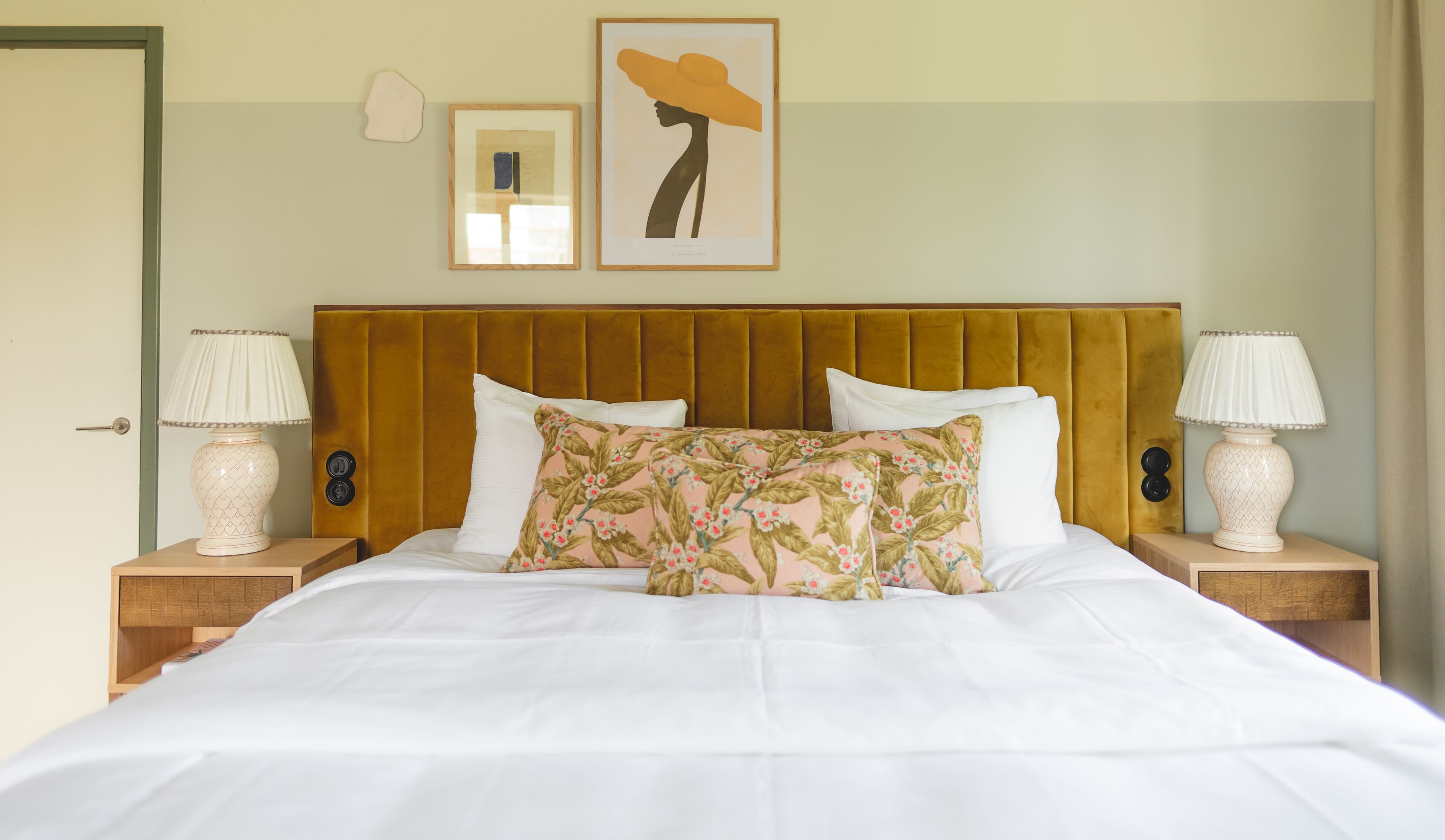 A neatly made bed with white linens, a floral pillow, and a large yellow hat artwork on the wall above.