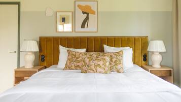 Un lit bien fait, avec du linge de lit blanc, un coussin à motifs floraux et, au-dessus, un grand tableau représentant un chapeau jaune.