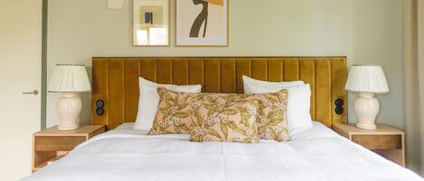 A neatly made bed with white linens, a floral pillow, and a large yellow hat artwork on the wall above.