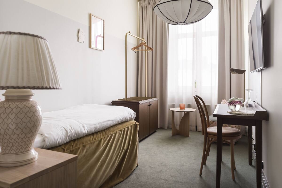 Habitación de hotel con cama, mesita de noche, lámpara, escritorio, silla y televisor montado en la pared.