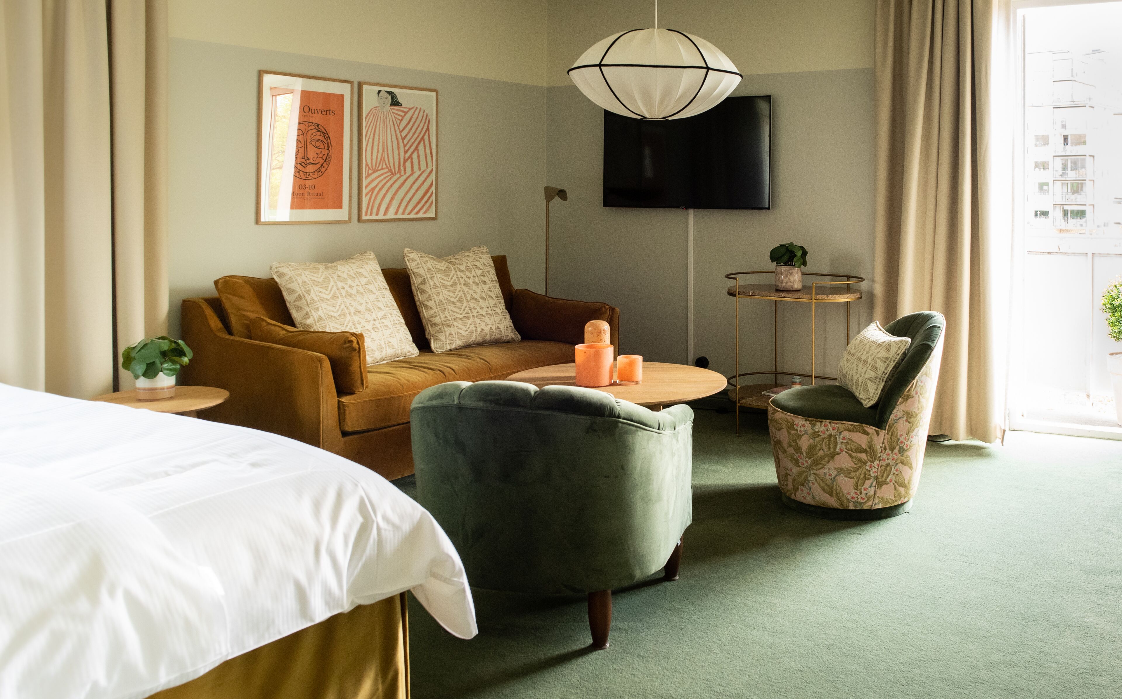 A hotel room with a bed, a sofa, a green armchair, a small table with candles, a TV mounted on the wall, and a window with curtains.