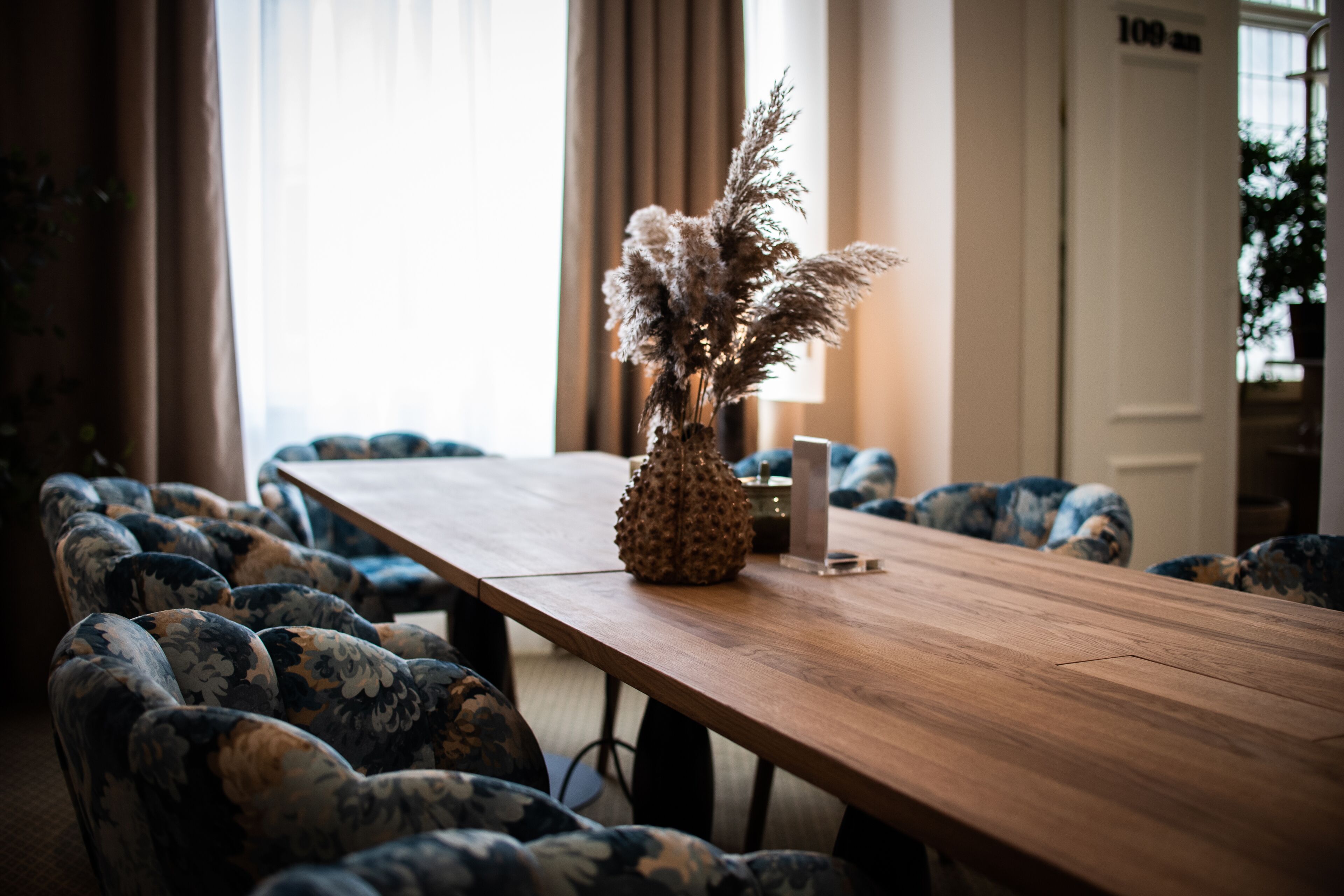 A conference room with a long wooden table, floral-patterned chairs, and a vase with dried flowers.