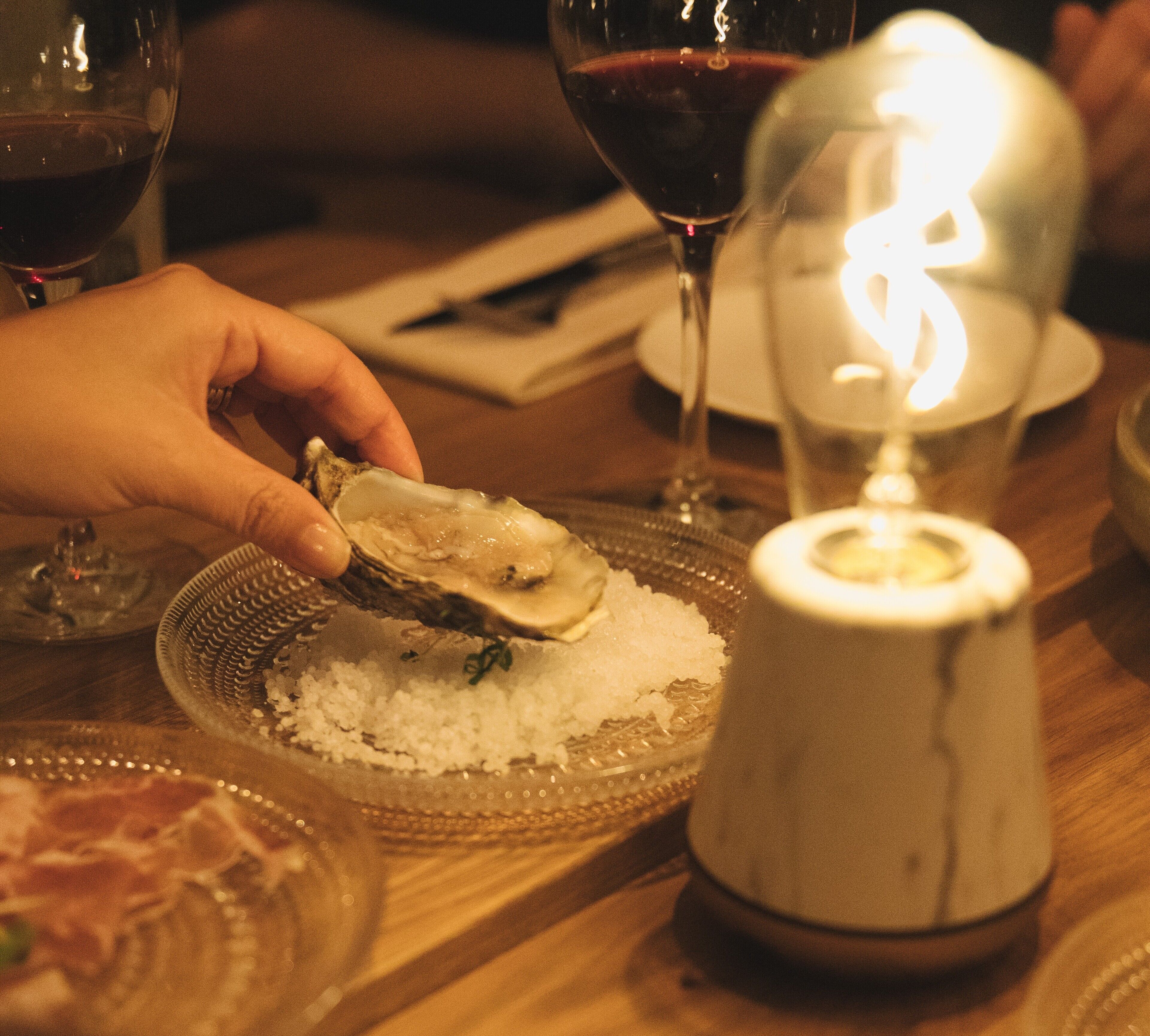 A dining table with a plate of oysters on ice, a glass of red wine, and a lit candle.