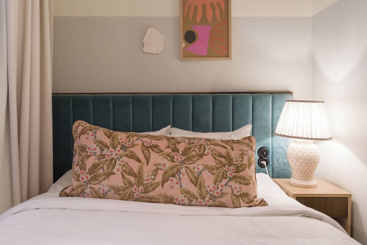 Una cama bien hecha con un cojín floral, un cabecero color turquesa, una lámpara en una mesita de noche y una obra de arte en la pared.