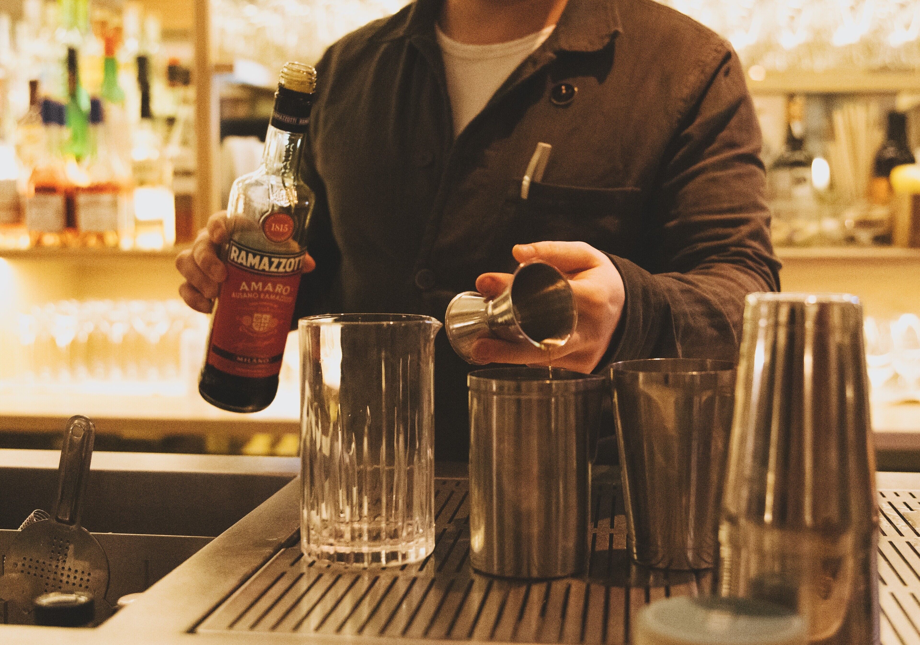 Un bar con una persona sirviendo una bebida en un vaso, una botella de licor y varios cocteleras sobre la barra.
