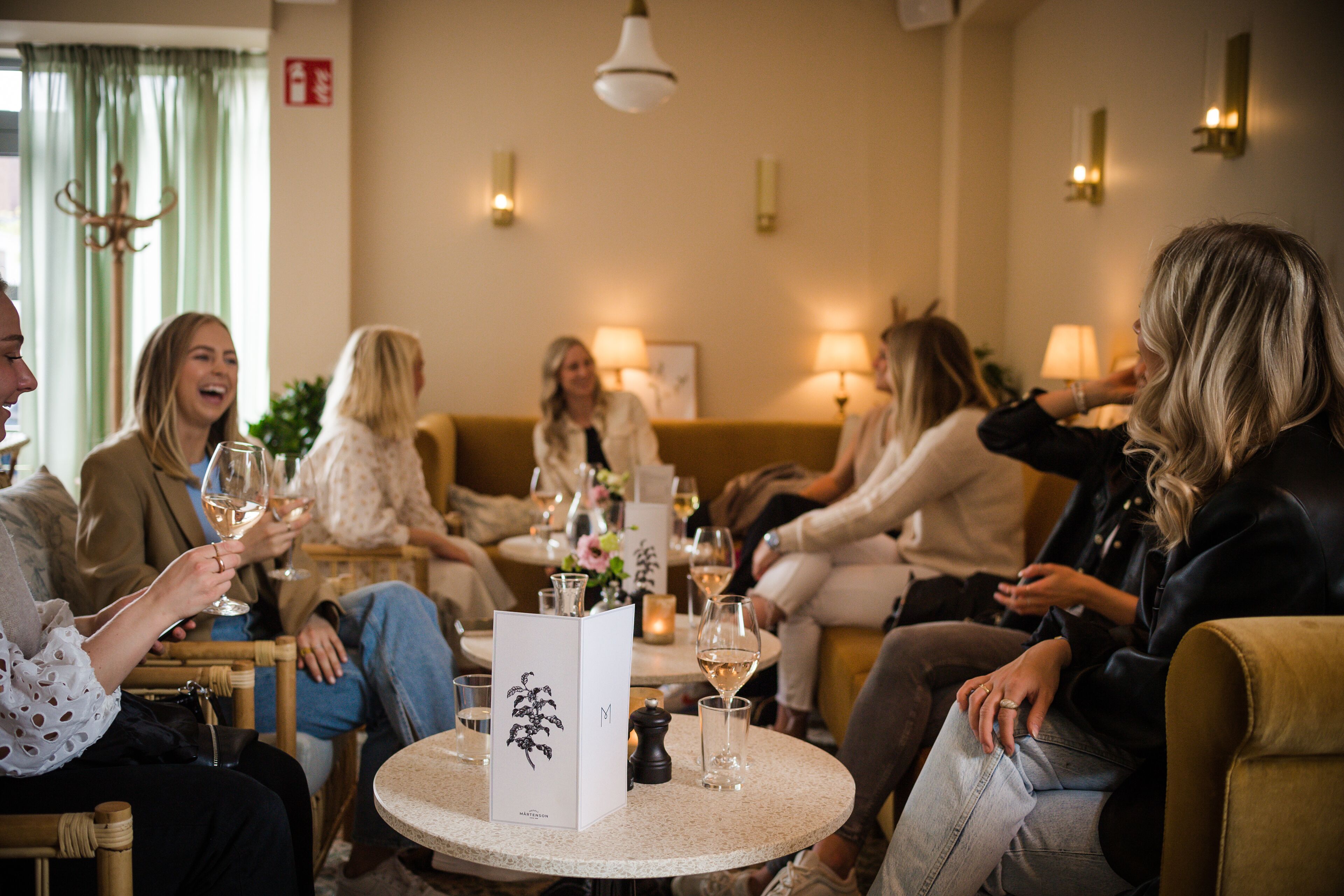 Un grupo de personas sentadas alrededor de una mesa redonda en una habitación bien iluminada, con un candelabro, una cortina verde y lámparas de pared.