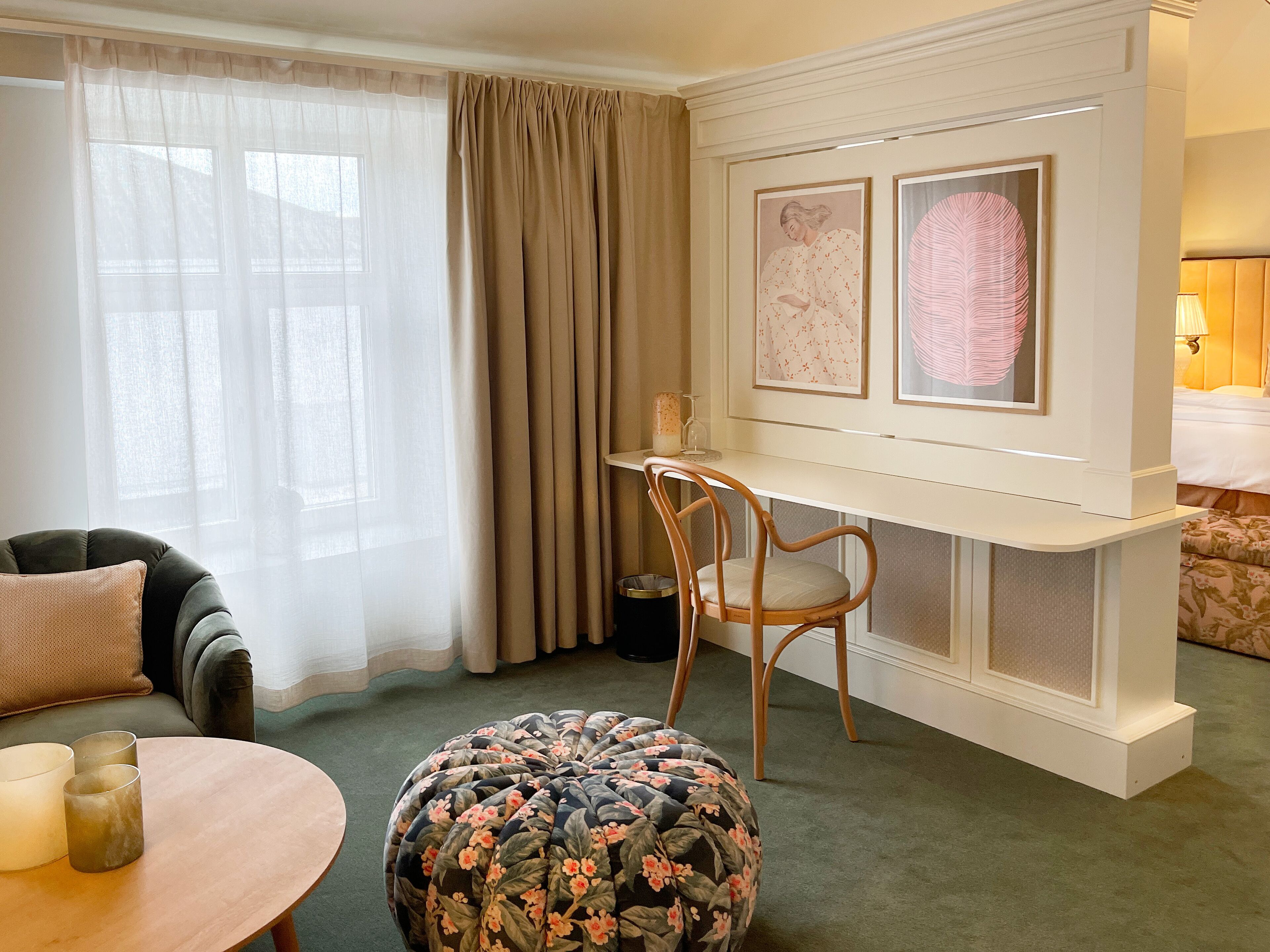 A well-lit room with a round wooden table, a floral ottoman, a wooden chair, a desk with a chair, and two framed artworks on the wall.