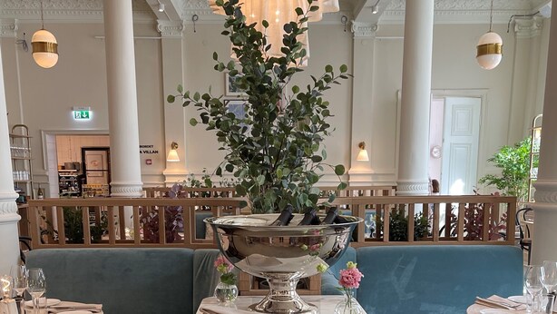A restaurant with a blue upholstered bench, a wooden table with a vase of flowers, and a large decorative mirror. - Elite Hotel Mårtenson (Halmstad)
