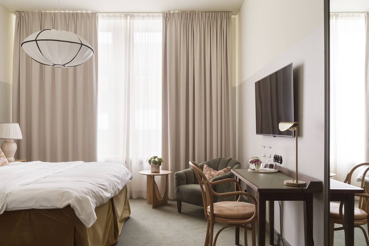 Una habitación de hotel con cama, escritorio, silla, televisor y un ventanal con cortinas.
