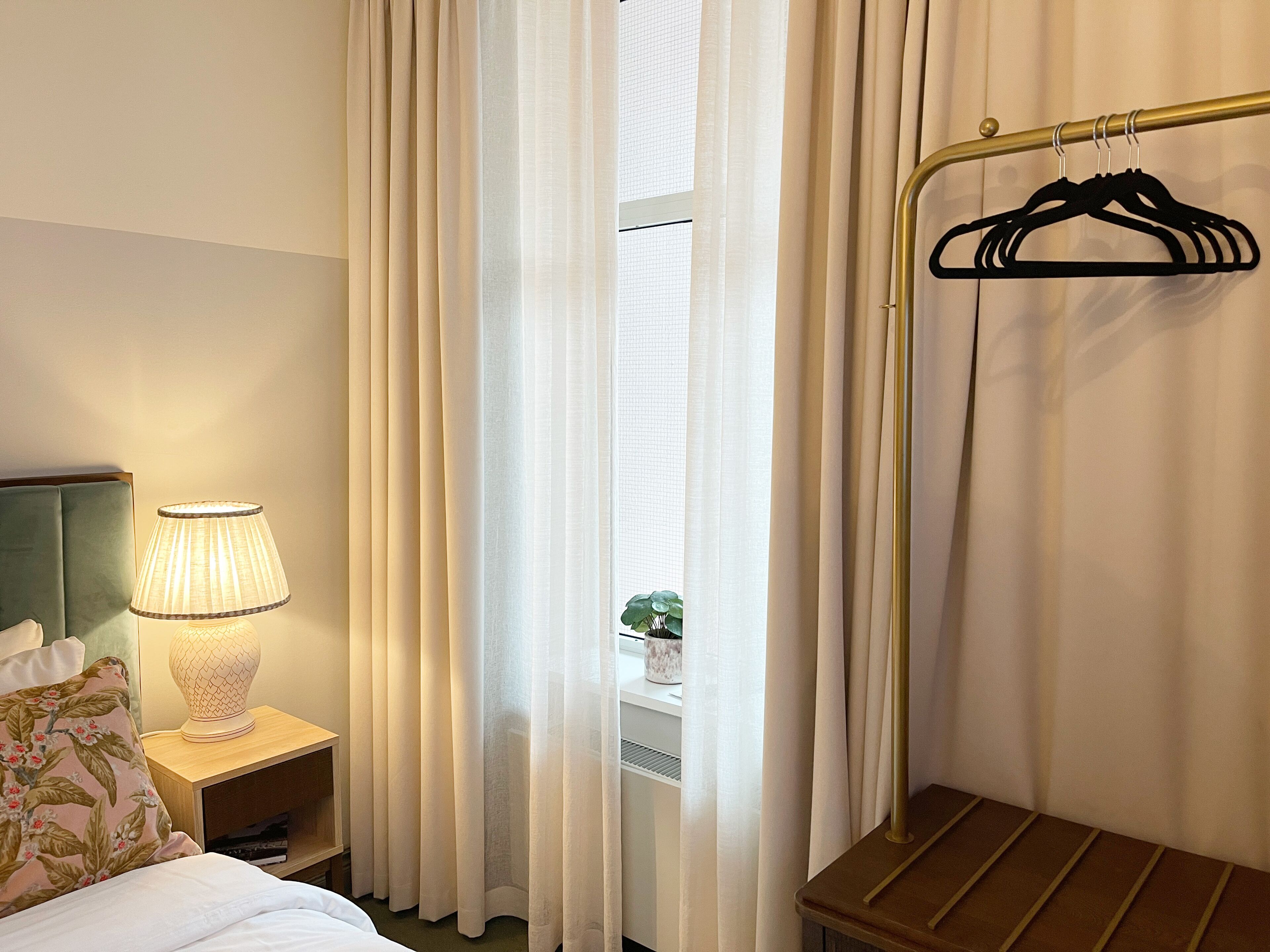 A hotel room with a bed, a nightstand with a lamp, a wooden bench, and a clothing rack.