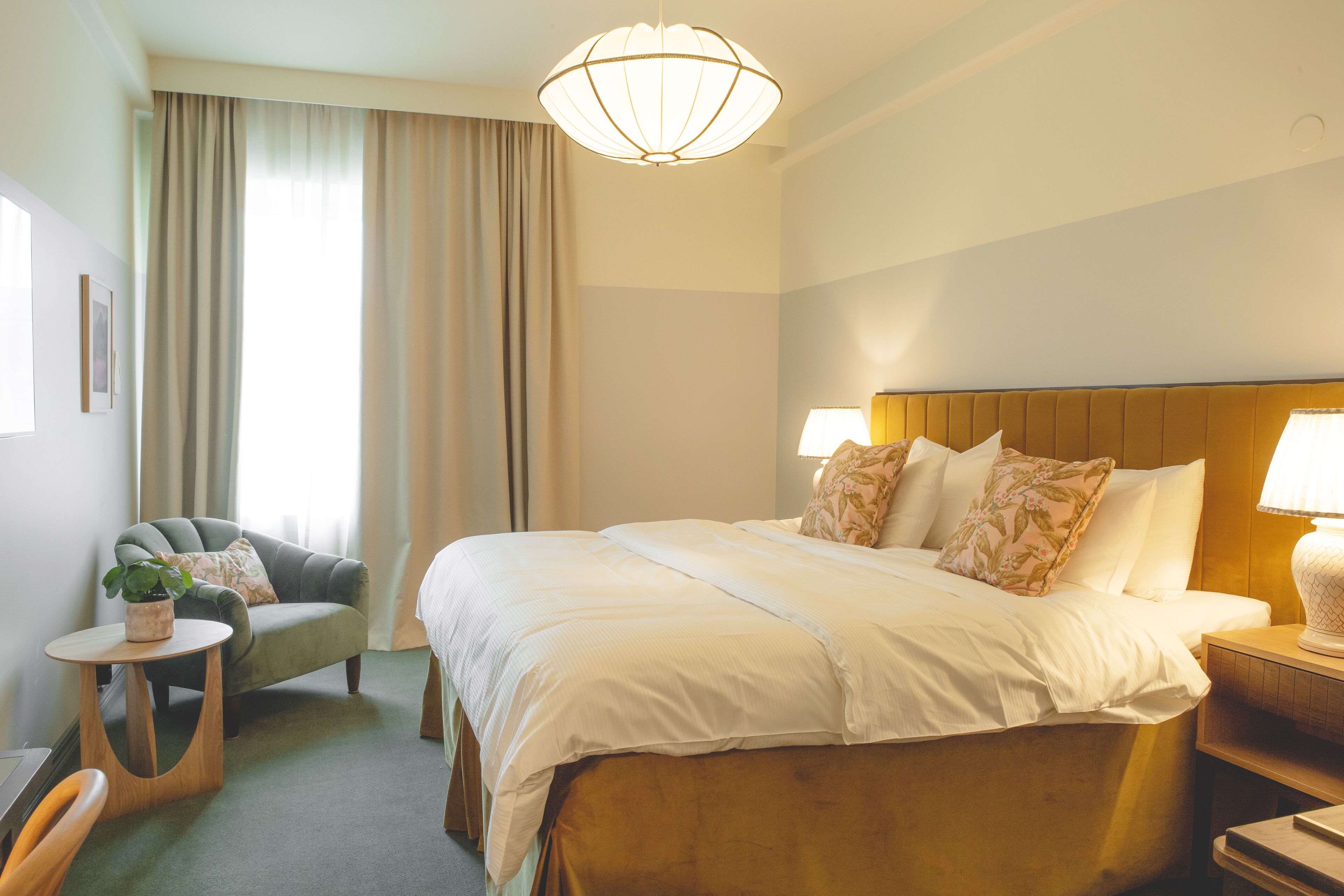 A hotel room with a large bed, a chair, a small table with a plant, and a nightstand with a lamp.