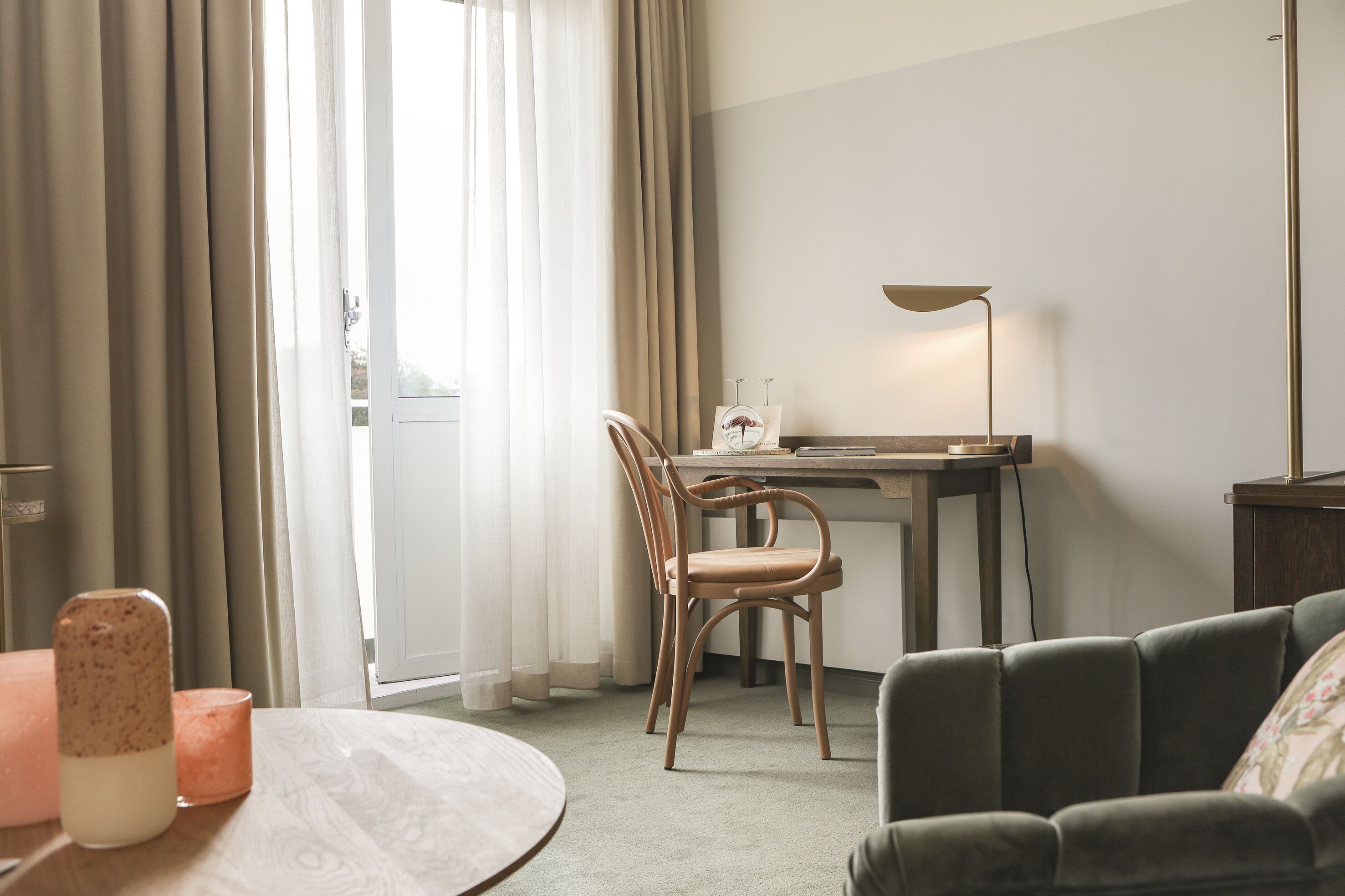 Quarto de hotel com mesa de jantar, cadeira, sofá, escrivaninha com luminária e janela com cortinas.