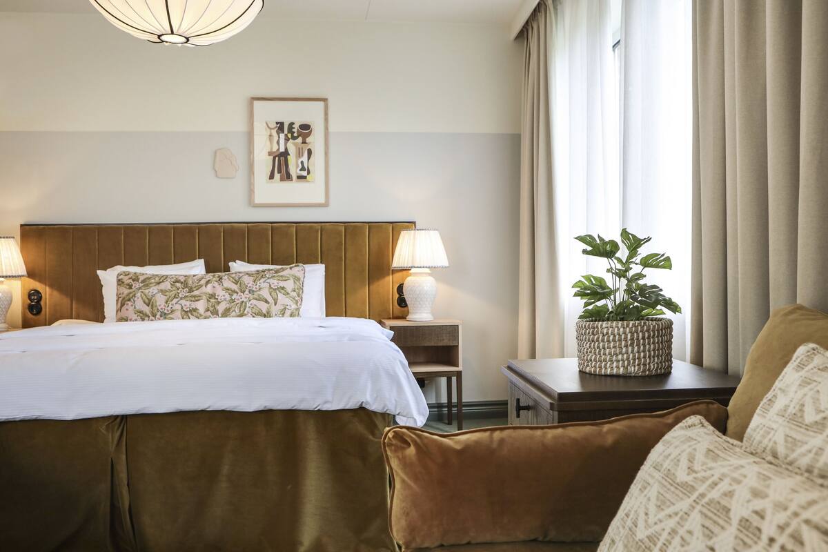 Habitación de hotel con una cama grande, dos lámparas de noche, una mesita de noche con una planta y una ventana con cortinas.