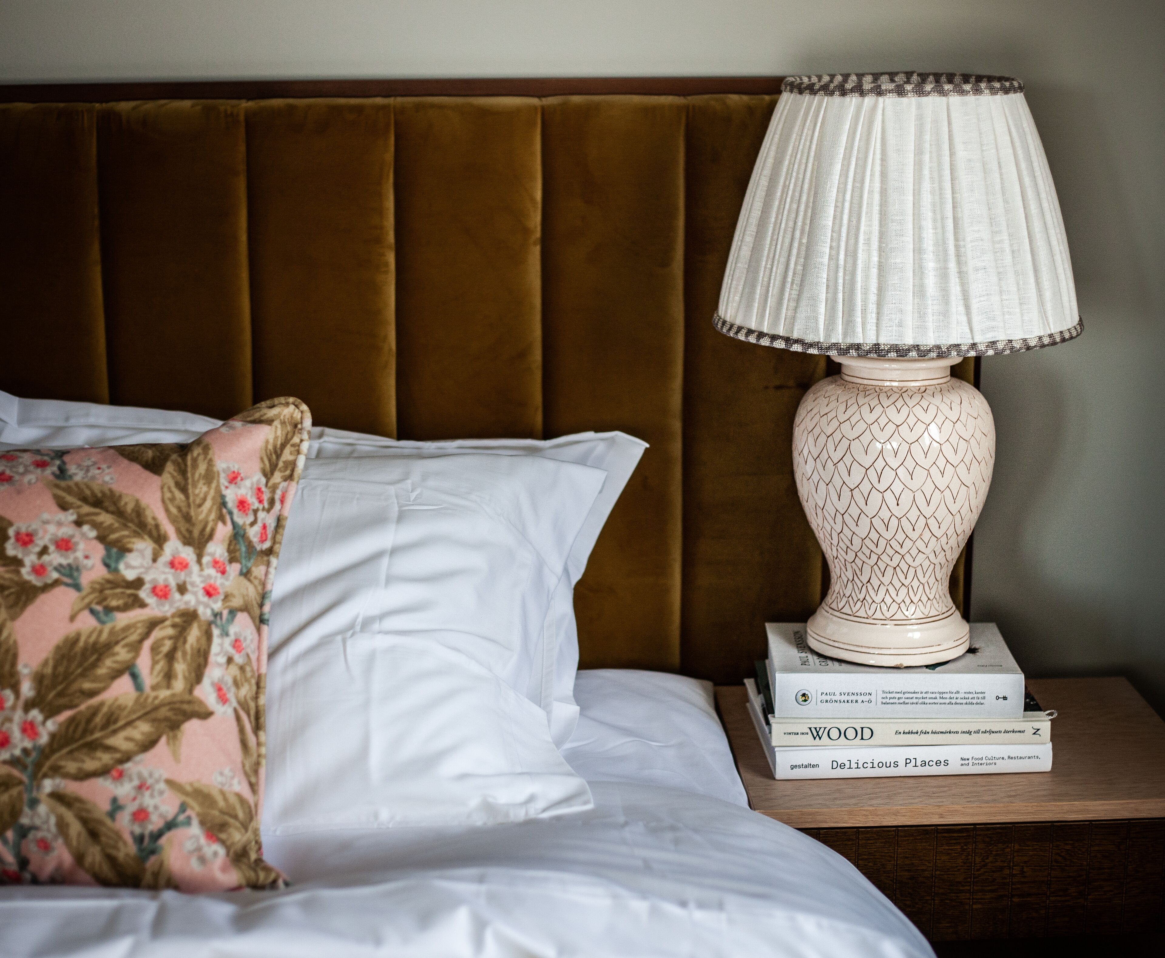 A neatly made bed with white linens and a floral pillow, a white lamp with a patterned shade, and a stack of books on a wooden nightstand.