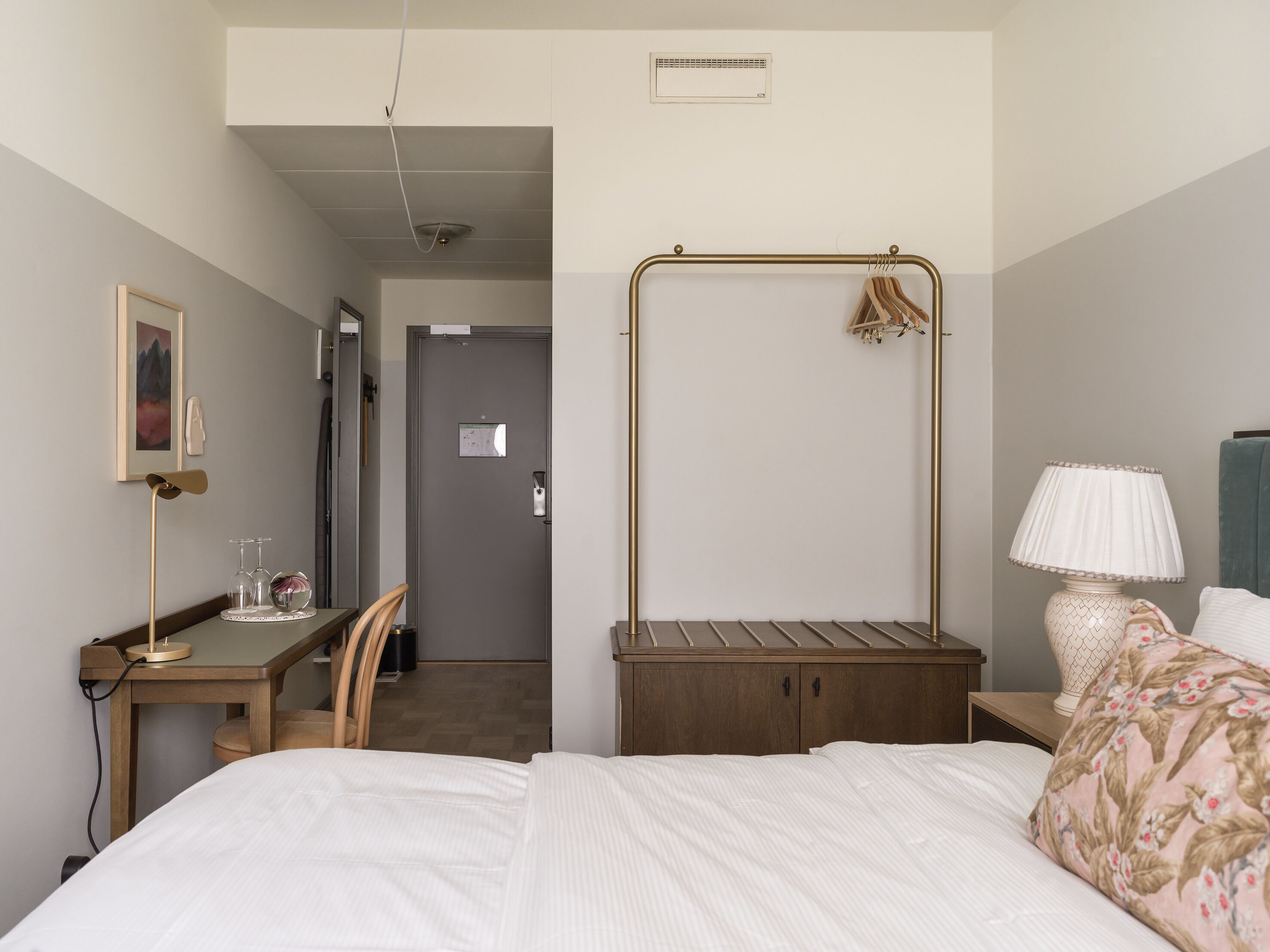 A hotel room with a bed, a desk, a chair, a hanging coat rack, a bedside lamp, and a door leading to a hallway.