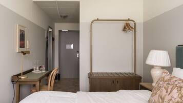 A hotel room with a bed, a desk, a chair, a hanging coat rack, a bedside lamp, and a door leading to a hallway.