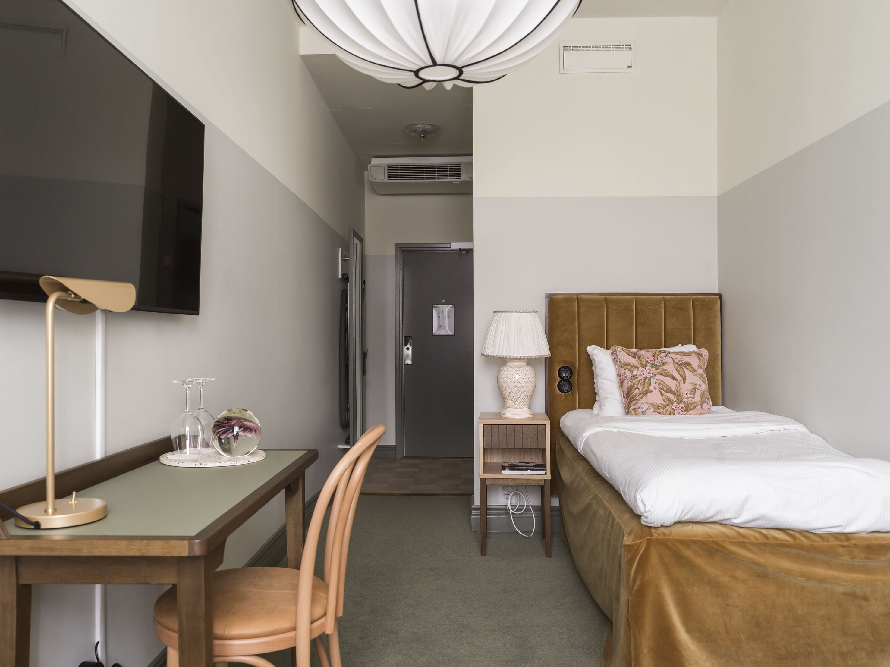 A hotel room with a bed, a desk, a chair, a TV, a lamp, and an air conditioning unit.