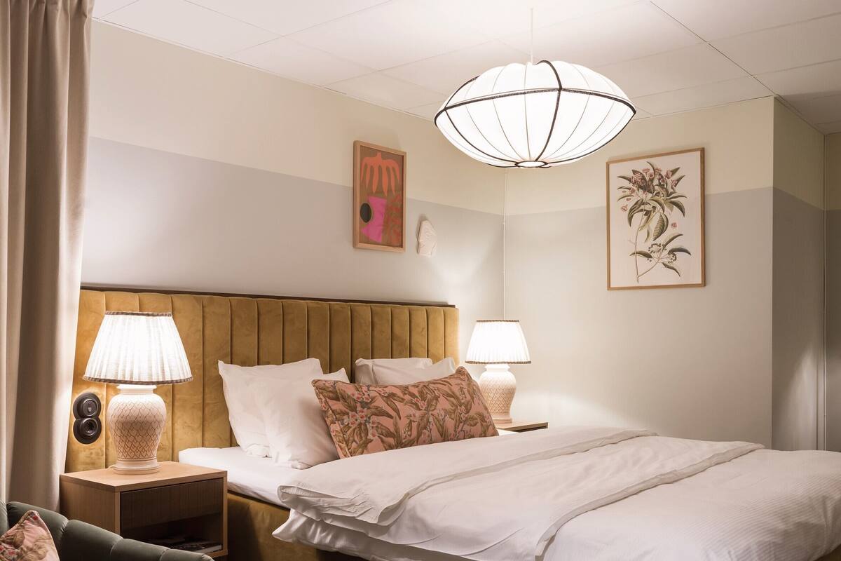 Habitación de hotel con una cama grande, dos lámparas de noche, una mesita de noche, una silla y obras de arte en las paredes.