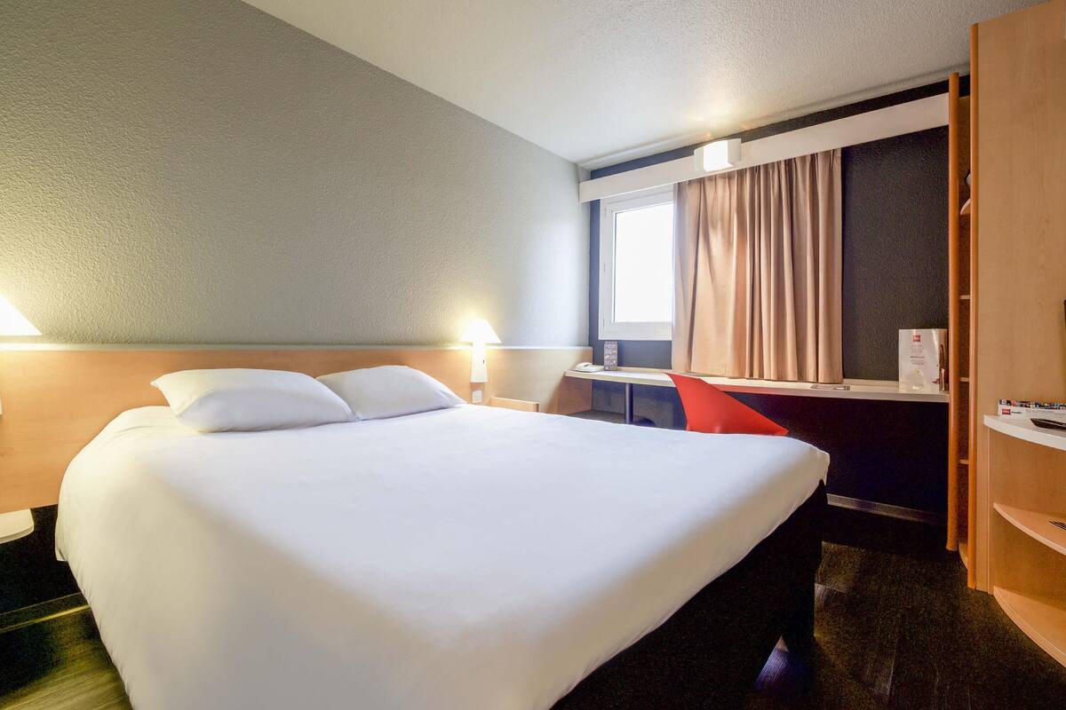Hotellihuone, jossa on suuri sänky, työpöytä ja tuoli. Huoneessa on ikkuna, jossa on verhot, sekä lamppu ja hylly, jossa on erilaisia esineitä.
