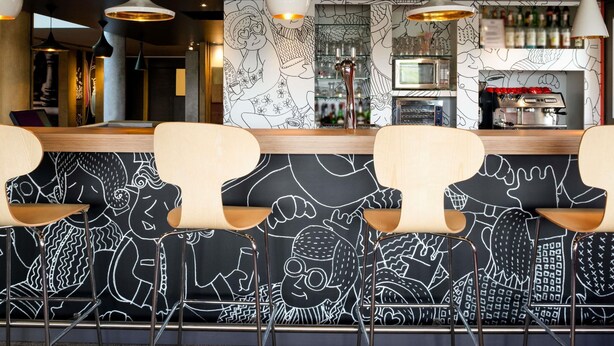 A bar area with a counter, bar stools, and a wall with a black and white mural. - ibis Pau Lescar (Lescar)