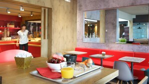 A breakfast setup with a tray of food, a bowl of fruit, a glass of orange juice, and a napkin on a wooden table in a restaurant with red seating and a mirror on the wall. - ibis Pau Lescar (Lescar)