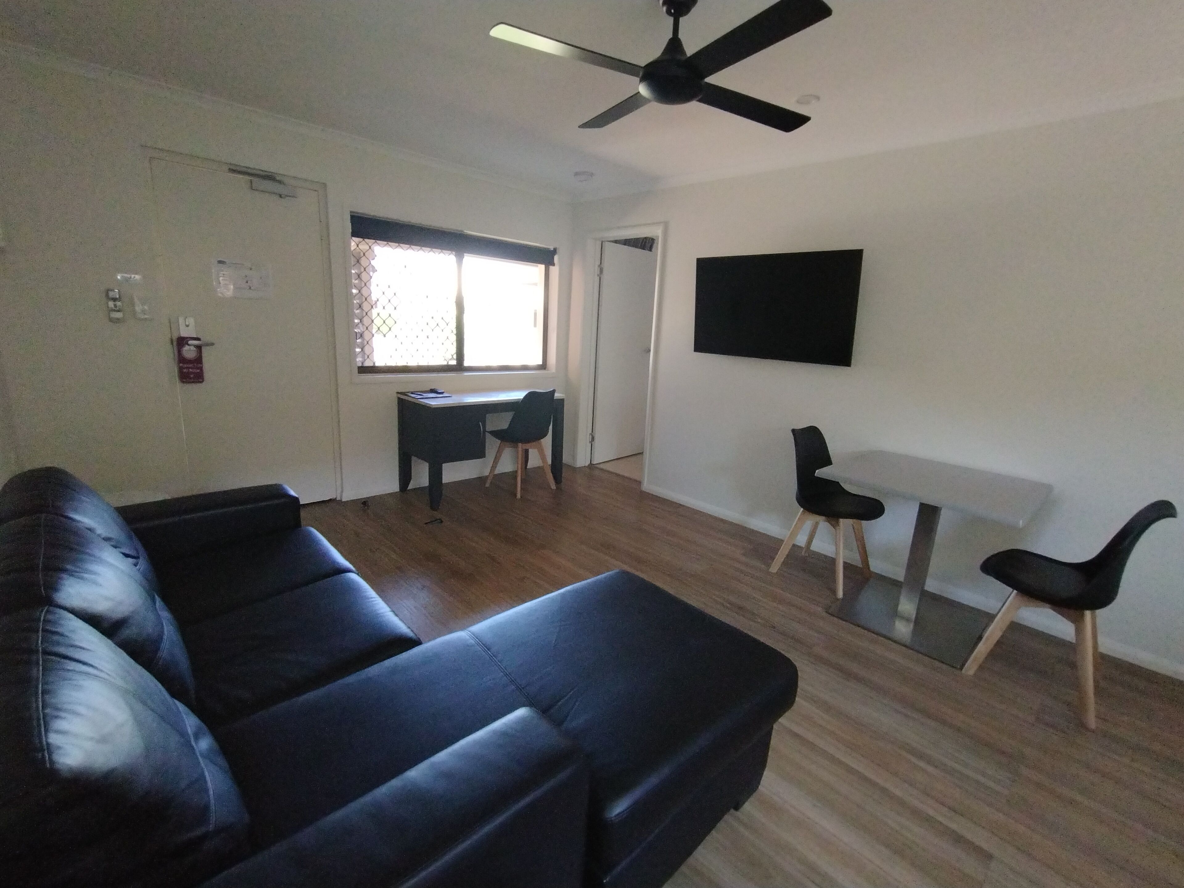 A living room with a black leather sofa, a dining table with chairs, a TV mounted on the wall, and a ceiling fan.