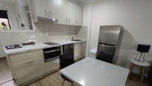 Una cocina moderna con horno empotrado, microondas y lavavajillas. Hay una mesa de comedor con sillas y una mesita auxiliar con una lámpara.
