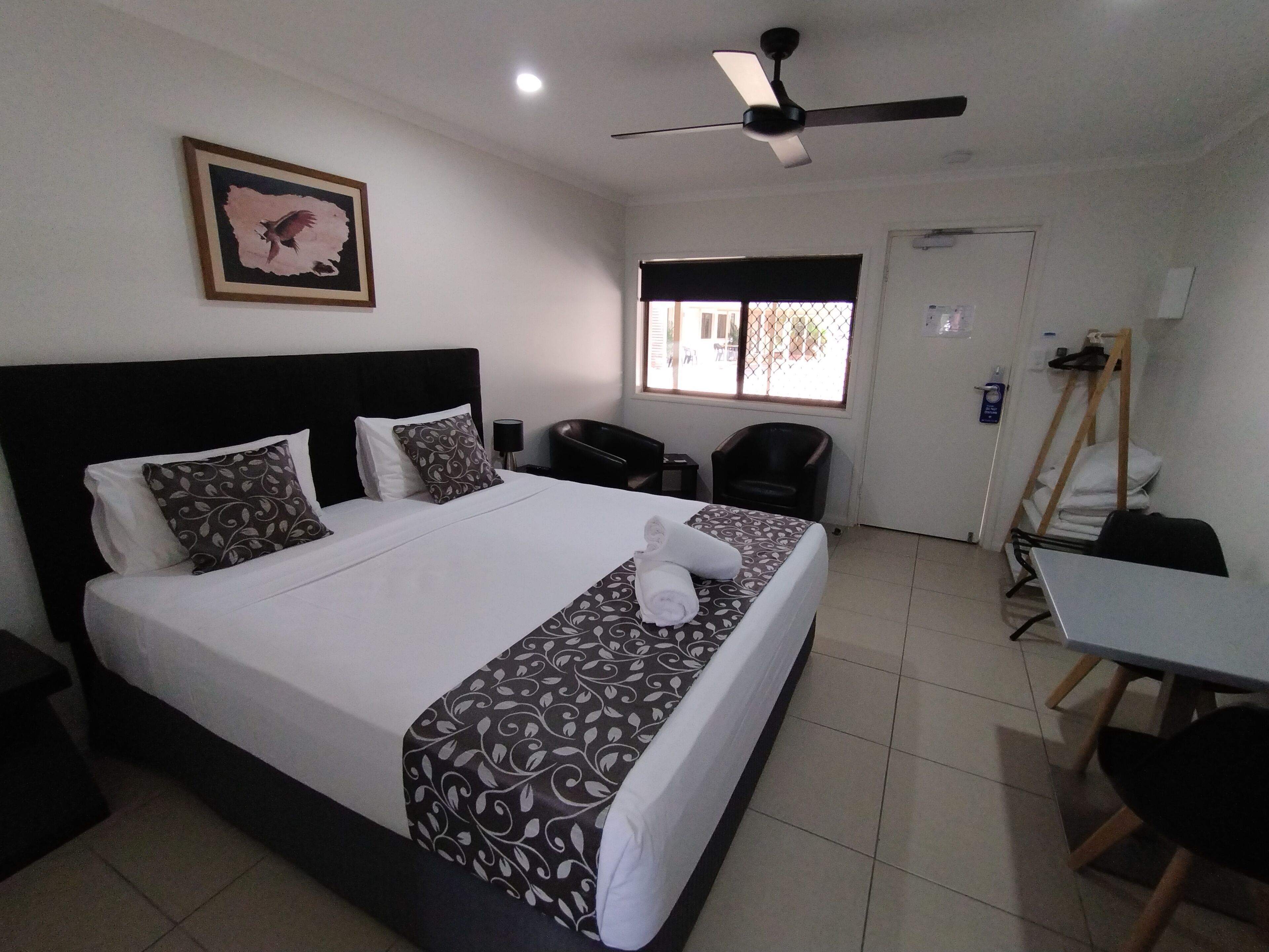 A hotel room with a bed, a desk, a chair, a ceiling fan, and a window with a view of the outside.