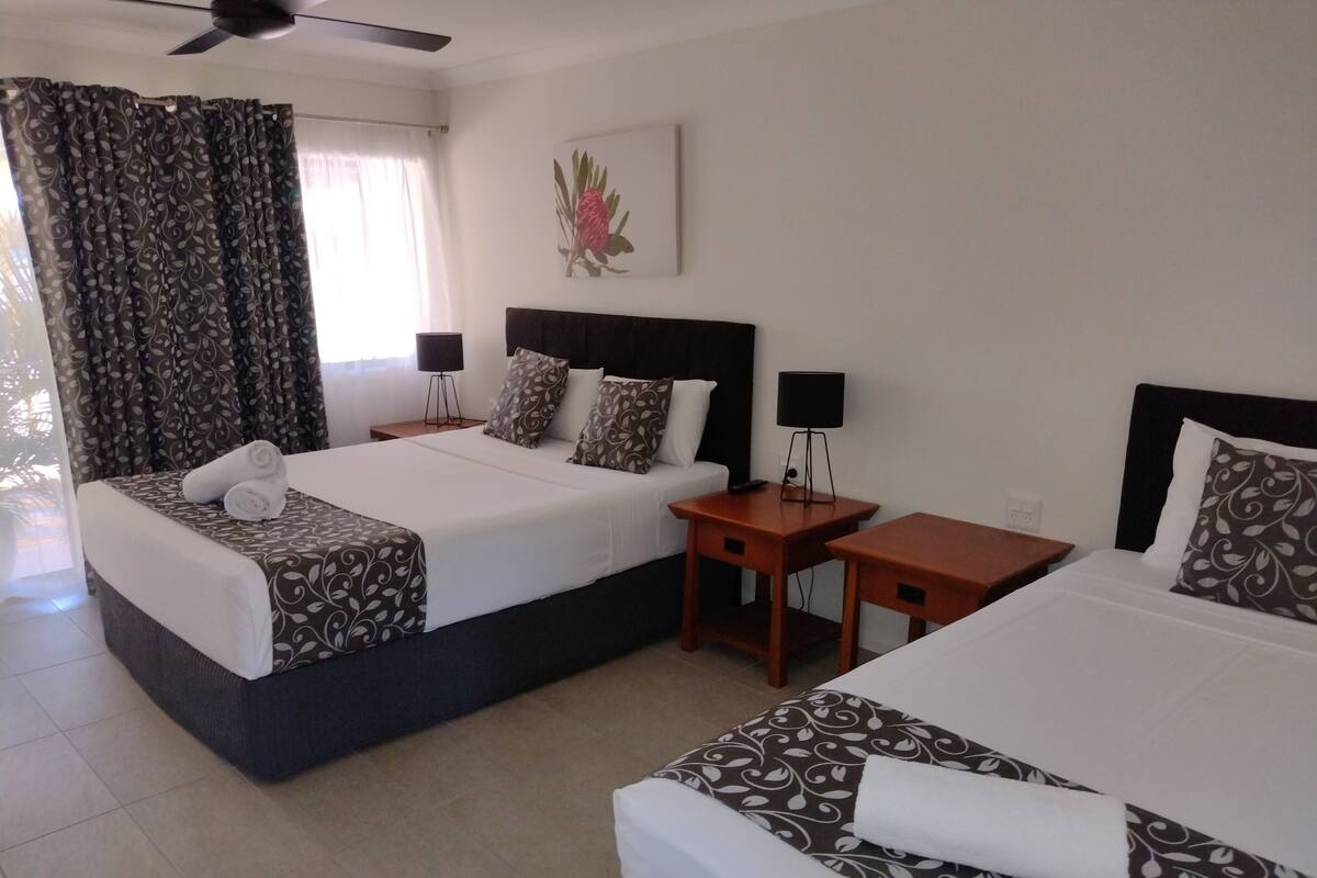 A hotel room with two beds, a ceiling fan, a window with curtains, a bedside table with a lamp, and a wall-mounted artwork.