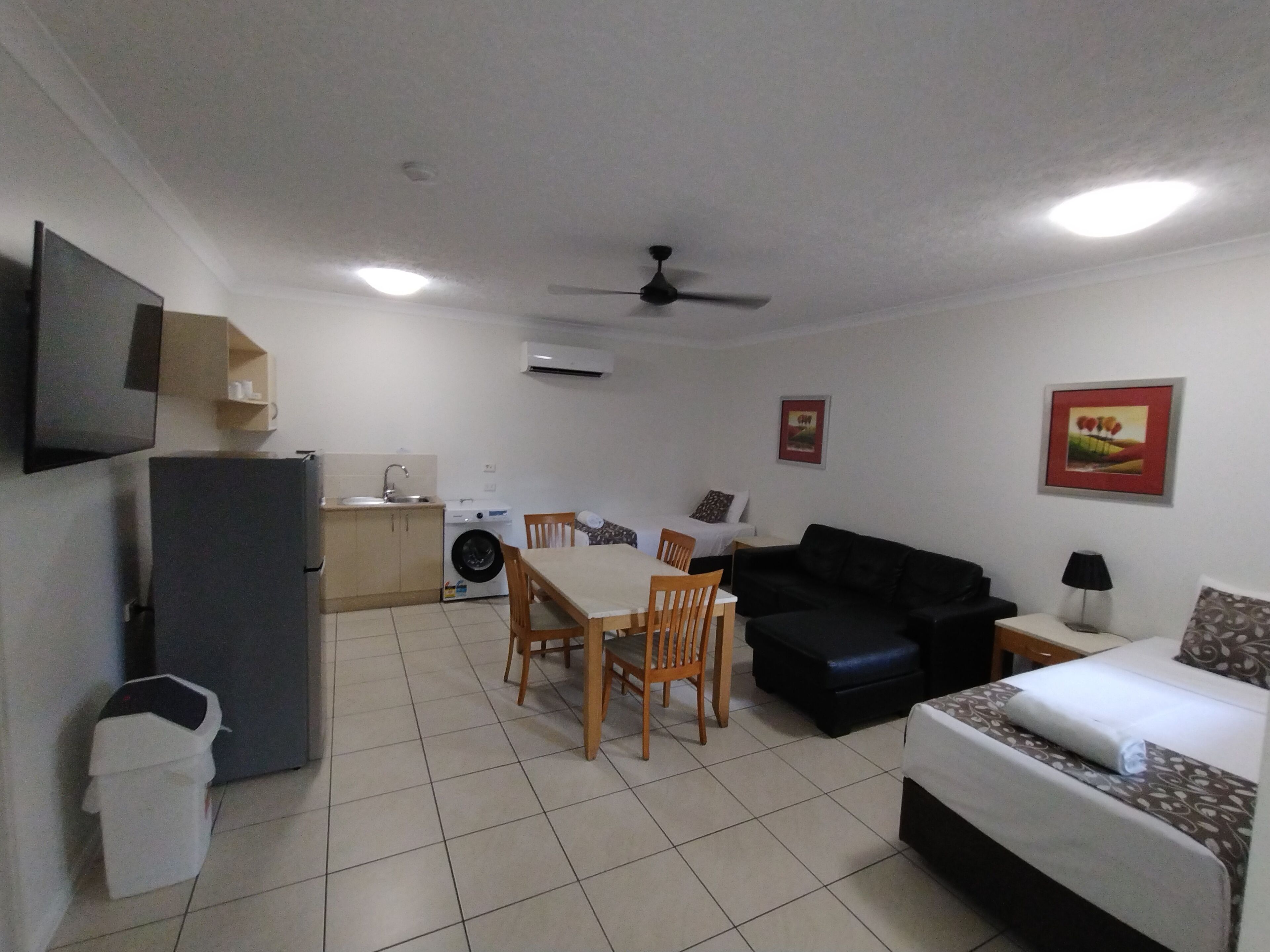 A compact living space with a dining area, a sofa, a bed, a washing machine, and a TV mounted on the wall.