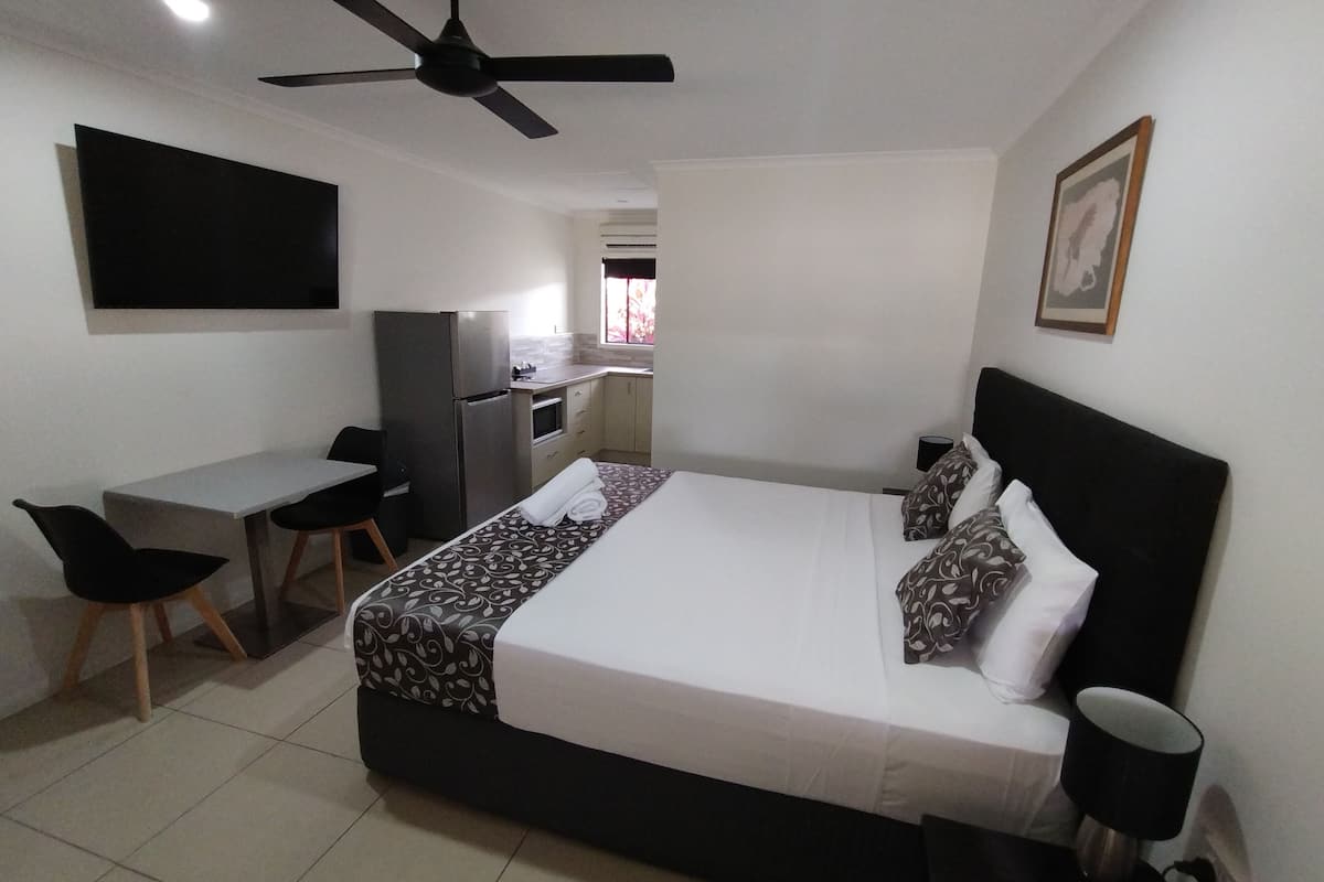 A bedroom with a bed, a desk, a chair, a ceiling fan, and a kitchenette in the background.