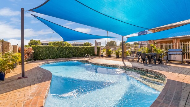 Una piscina con una toldo azul, zona de barbacoa y patio empedrado.