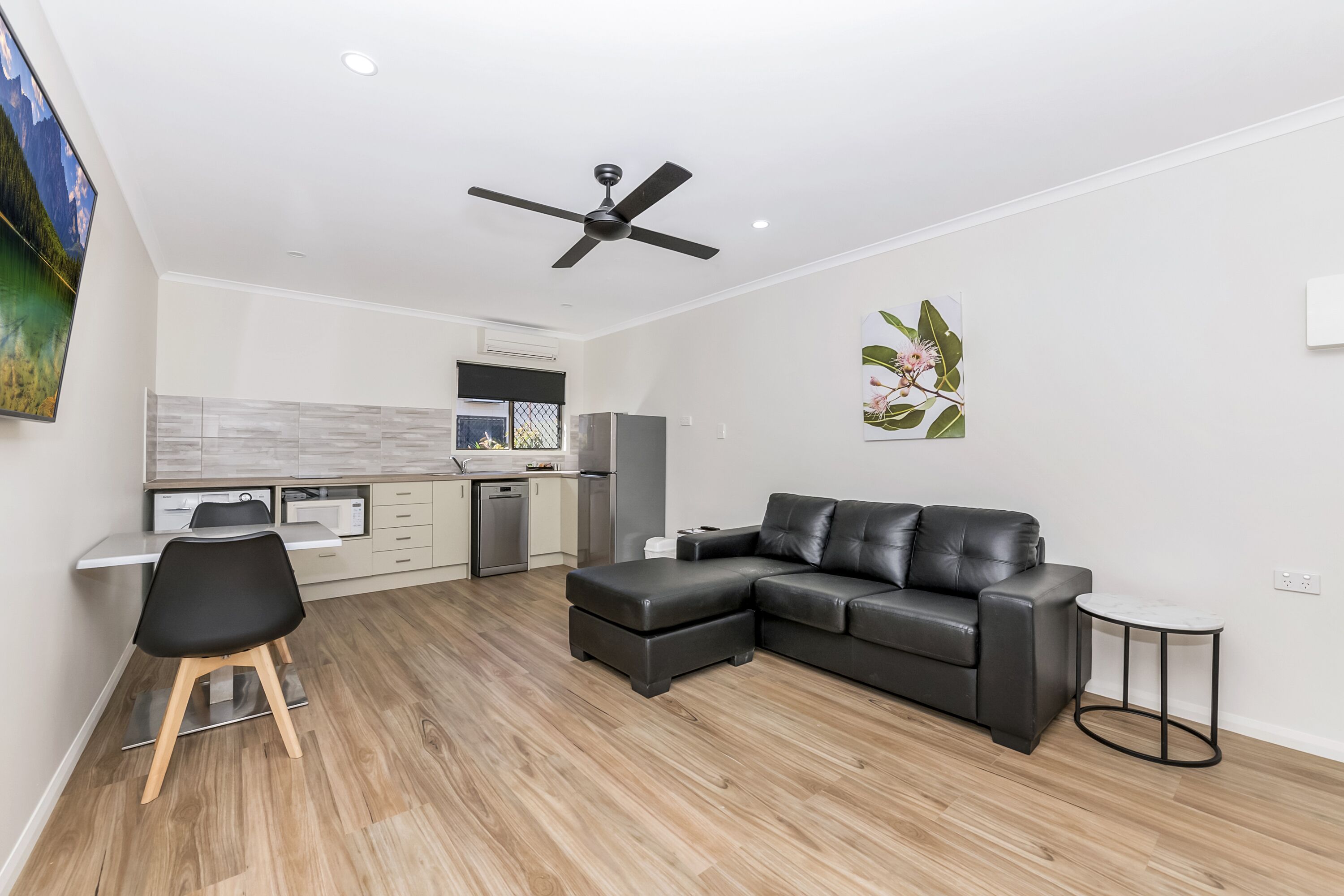 A modern living room with a black leather sofa, a dining area with a white table and chairs, and a kitchen in the background.
