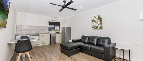 A modern living room with a black leather sofa, a dining area with a white table and chairs, and a kitchen in the background.