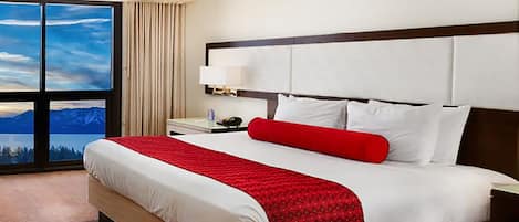 A hotel room with a large bed, a nightstand, a lamp, and a view of mountains through the window.