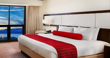 A hotel room with a large bed, a nightstand, a lamp, and a view of mountains through the window.